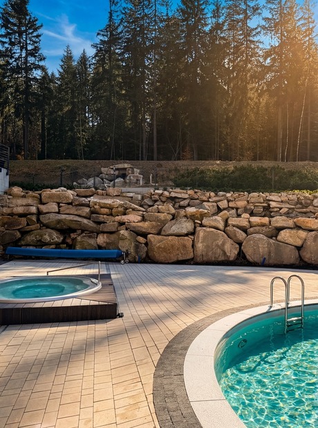 Rund utomhuspool med stege och intilliggande nedsänkt bubbelpool vid träklädd hotellterrass och stenmur framför skog.