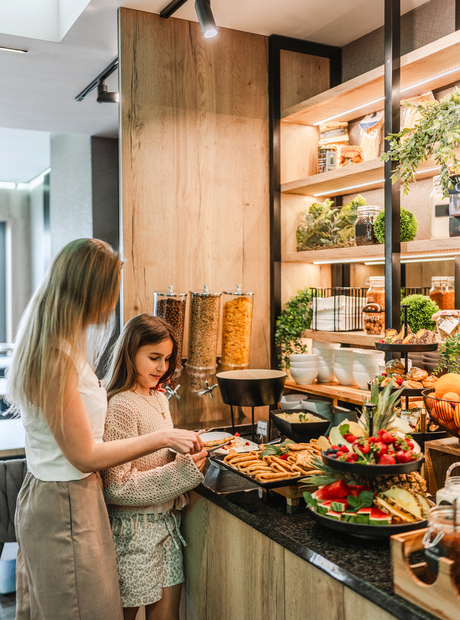 Matka z córką przy hotelowym bufecie śniadaniowym z ciepłymi drewnianymi akcentami, wybierają wypieki, świeże owoce i płatki.