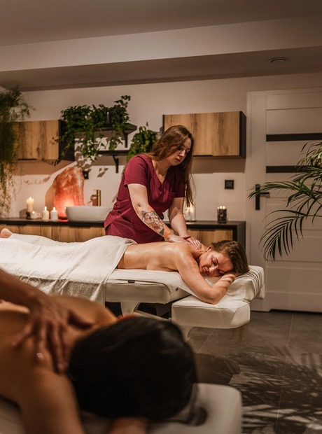 Serene spa treatment room with candles and plants; therapist massages guest on side-by-side treatment tables.