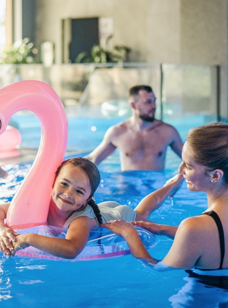 Innendørs hotellbasseng med blå LED-belysning; smilende jente på rosa oppblåsbar flamingoring holdt av kvinne, mann slapper av i bakgrunnen.