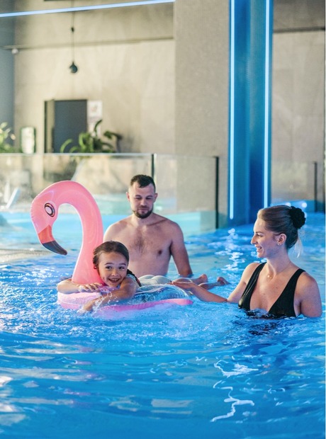 Family enjoying a modern indoor hotel pool; child on pink flamingo float, parents smiling while supervising in blue-lit spa pool.
