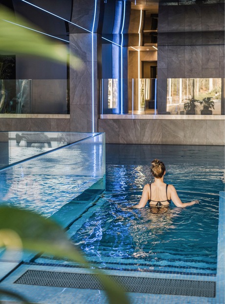 Gjest slapper av i moderne innendørs spa-basseng med glasskant, blå LED-lys og steinvegger