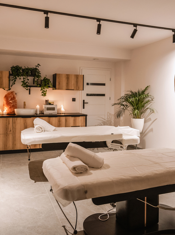 Ruhiger Spa-Raum mit zwei Massageliegen, aufgerollten Handtüchern, Kerzenlicht, Holzschränken, Pflanzen und großem runden Spiegel