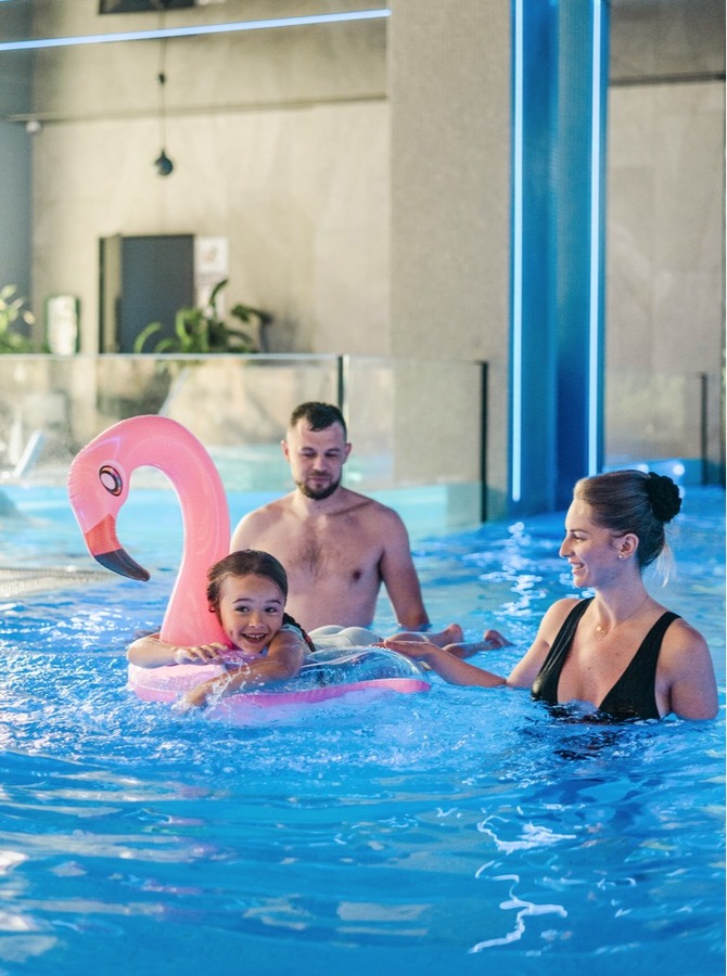 Familie i moderne innendørs hotellbasseng; barn på rosa flamingoring, foreldre smiler og passer på i blått opplyst spa.