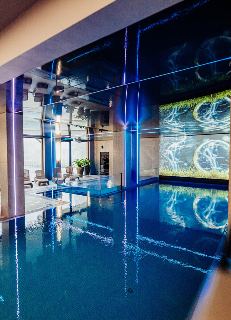 Innendørs hotellbasseng og boblebad med solsenger, blå LED-belysning og stor digital vannvegg