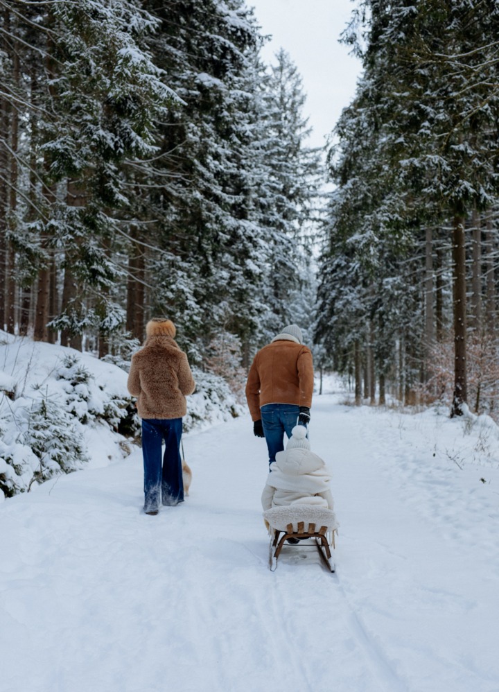 Par går på snødekt skogsti, drar treslede med innpakket barn, hund ved siden av, høye snødekte graner