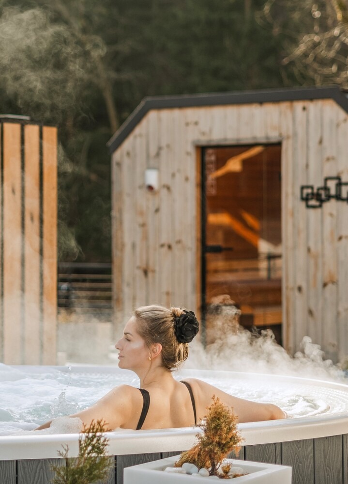 Två kvinnor kopplar av i en ångande utomhusbadtunna på hotellets spa, med träsaunor och skog i bakgrunden.