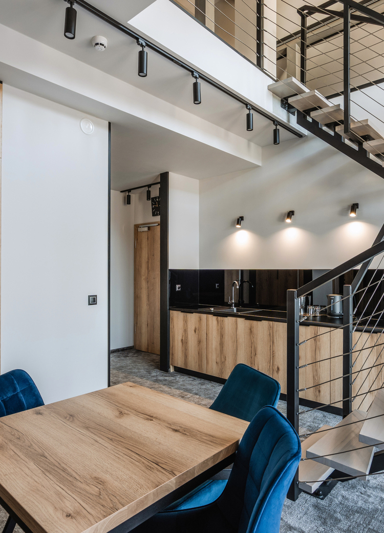 Moderní loft s dřevěnou kuchyňskou linkou, stolem s modrými židlemi, otevřeným kovovým schodištěm a útulnou ložnicovou nikou