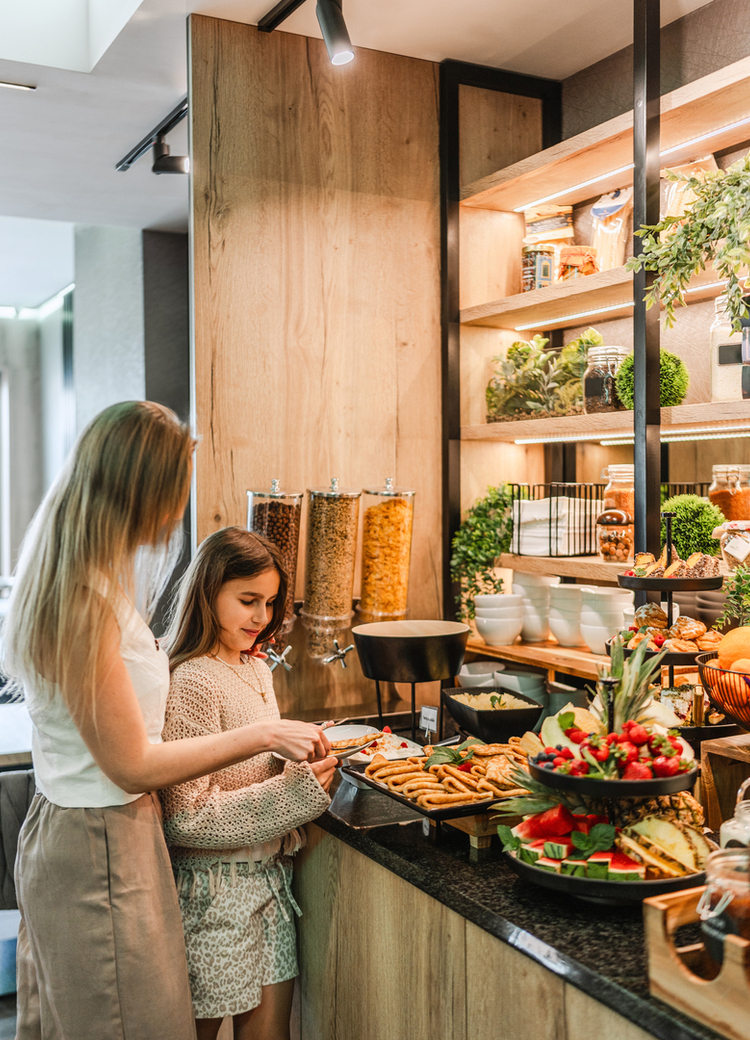 Mother and daughter at a hotel breakfast buffet choosing pastries, fruit and cereals by warm wood shelves and lit self‑service displays