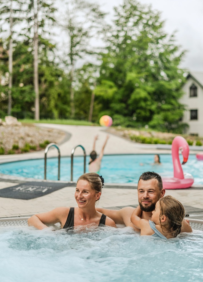 Family relaxing in outdoor hot tub beside resort pool with inflatable flamingo, trees and a villa in the background