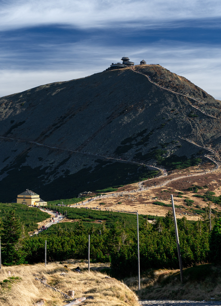 Berghütte und Aussichtsbau am Gipfel, geschwungener Wanderweg mit Wanderern, felsige Hänge und weites Himmelspanorama.