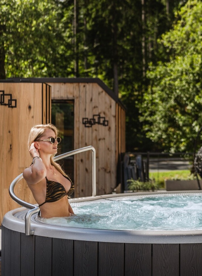 Bąbelkowe jacuzzi na hotelowym tarasie, kobieta w bikini i okularach, drewniane sauny i leśny widok