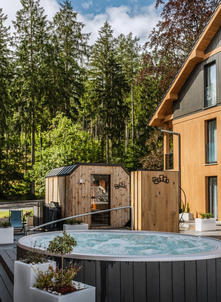 Venkovní vířivka a dřevěné sauny u moderního dřevěného hotelu, lesní prostředí, slunná spa terasa.