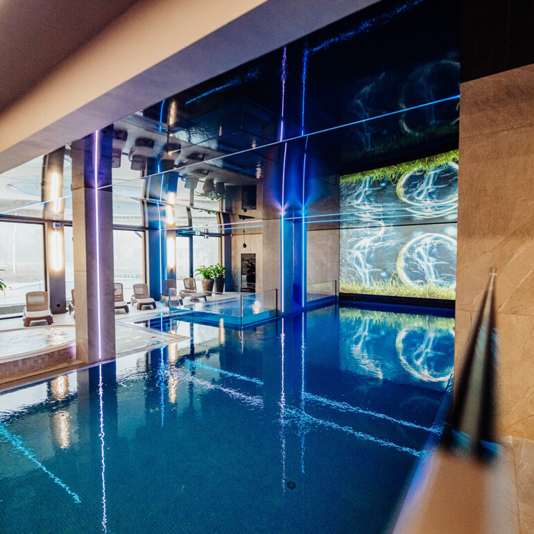 Innenpool und Whirlpool im Hotelspa mit Liegestühlen, blauer LED-Beleuchtung und großer digitaler Wasserprojektion