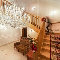 Foyer hotelu z kryształowym żyrandolem, drewnianymi schodami z czerwonym dywanem, ozdobną sofą i rozmazaną postacią