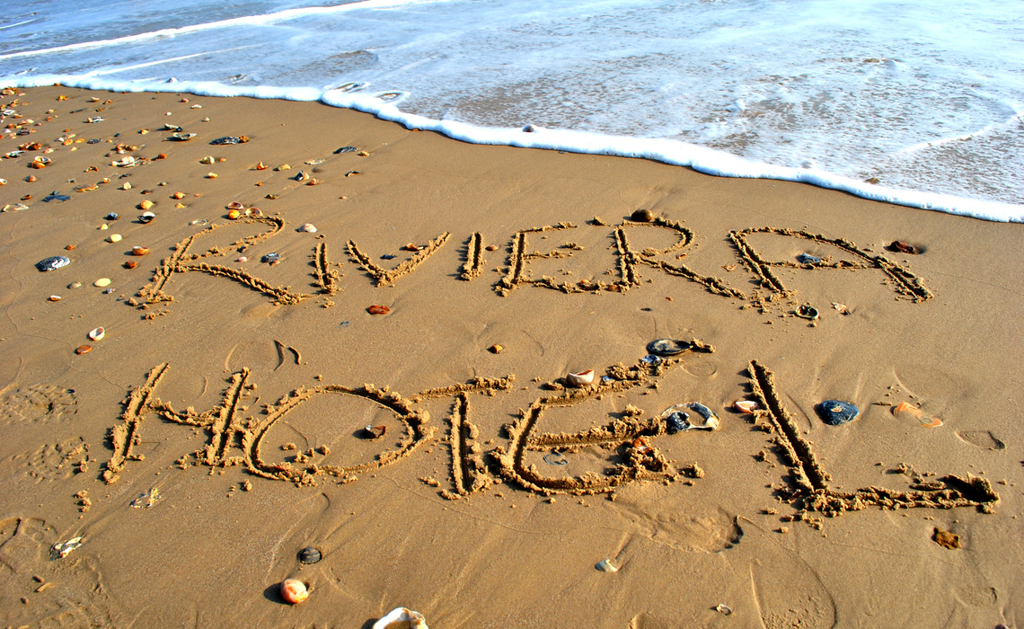 RIVIERA HOTEL written in sand on a shell-strewn beach as gentle surf laps the shoreline