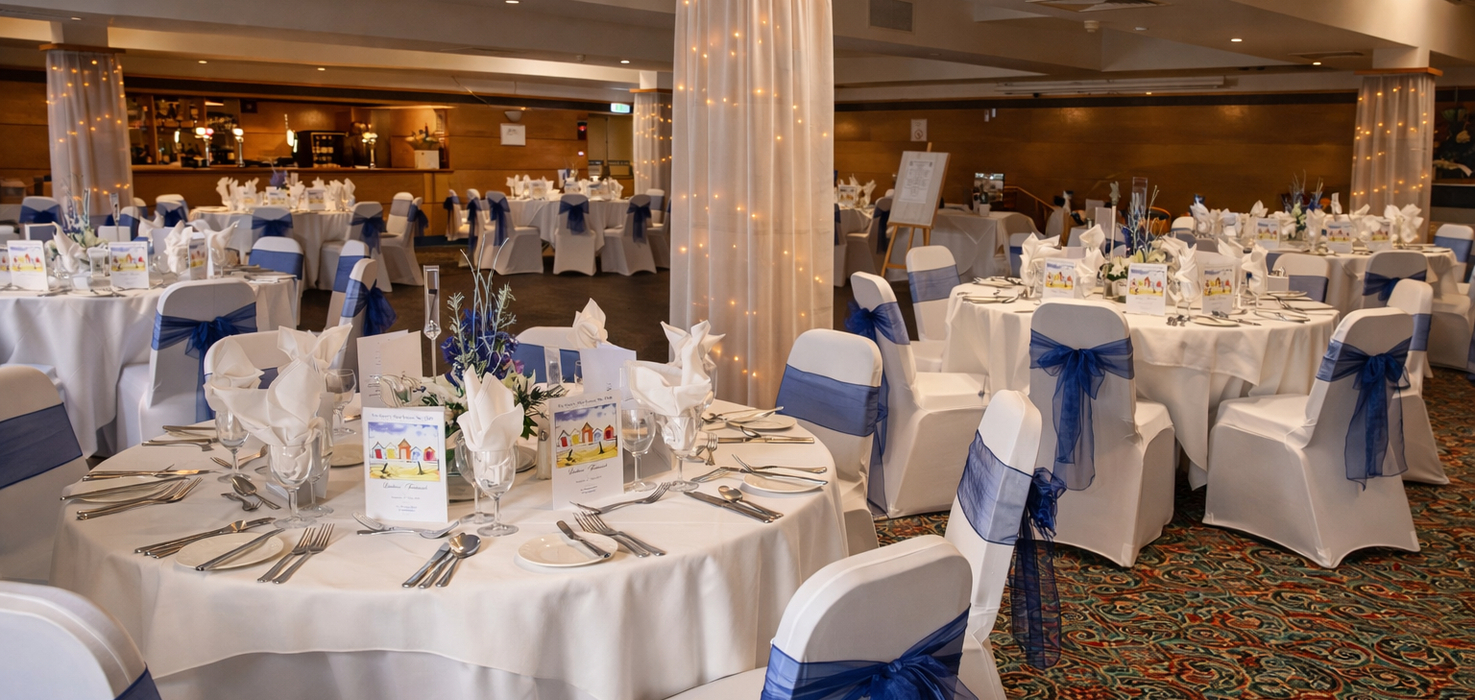 Elegant banquet hall set for wedding reception: round tables with white linens, navy-blue chair sashes, floral centerpieces and fairy-lit columns.