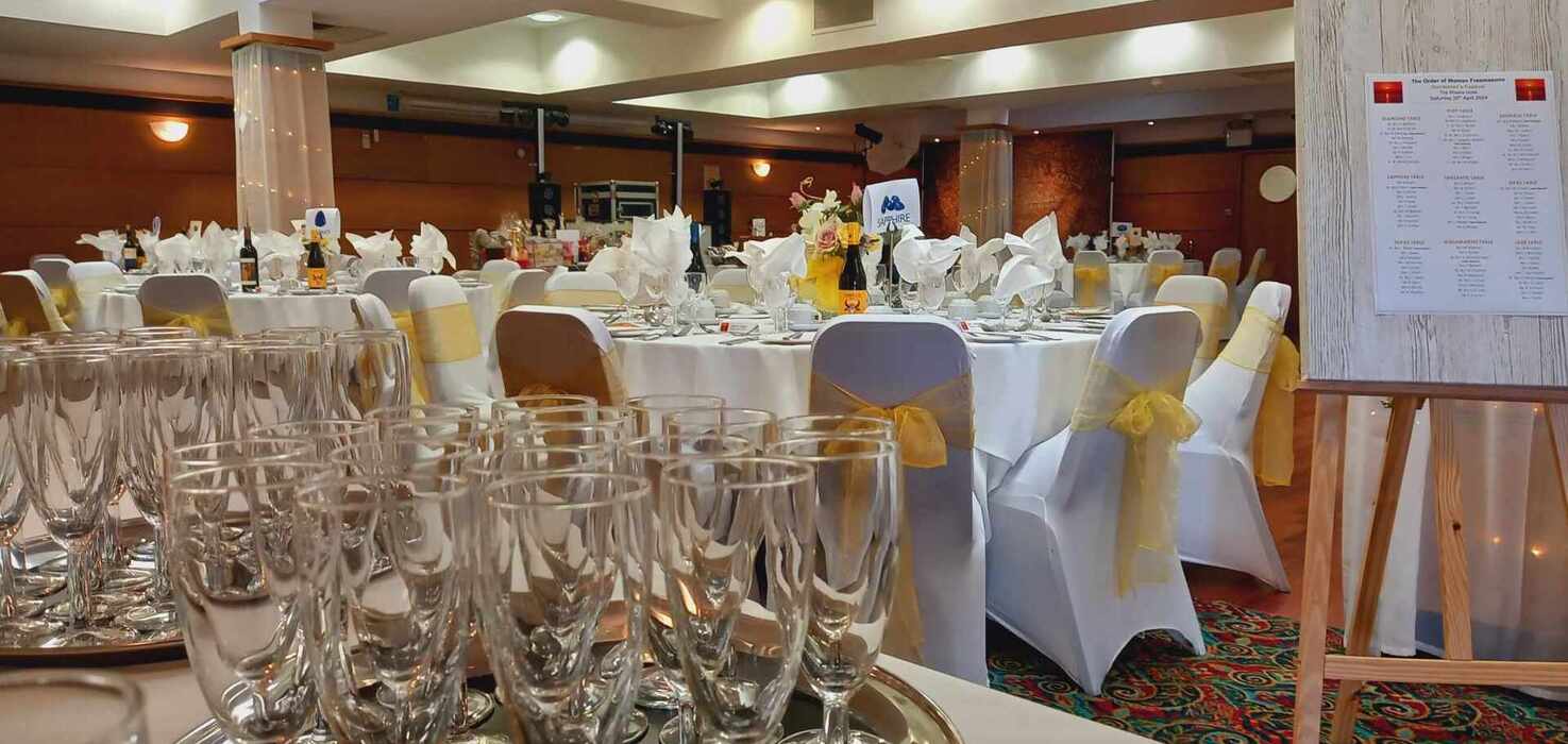 Banquet hall set for reception: round tables with white linens, napkins, floral centerpieces, wine bottles, chairs with yellow bows, glassware.