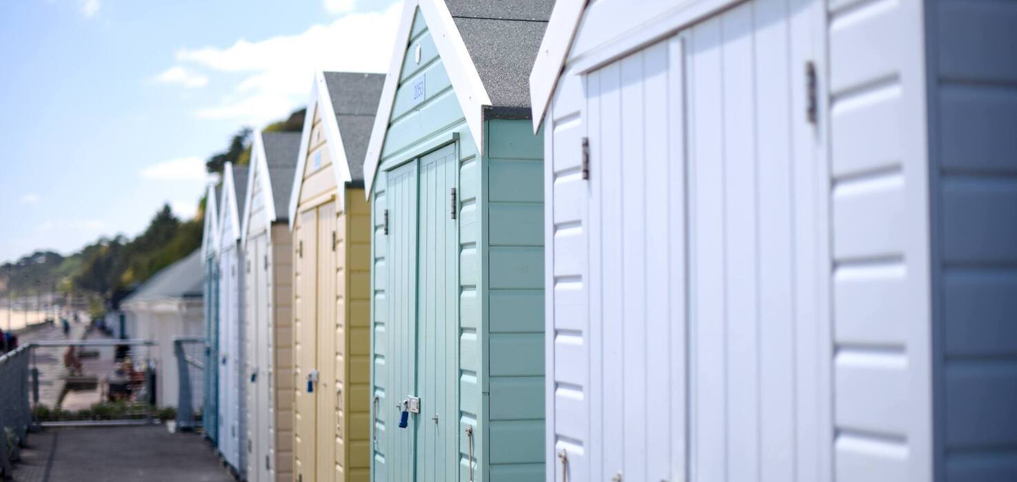 Row of pastel beach huts with pitched roofs and padlocks lining a sunny seaside promenade, blurred beach and trees beyond.