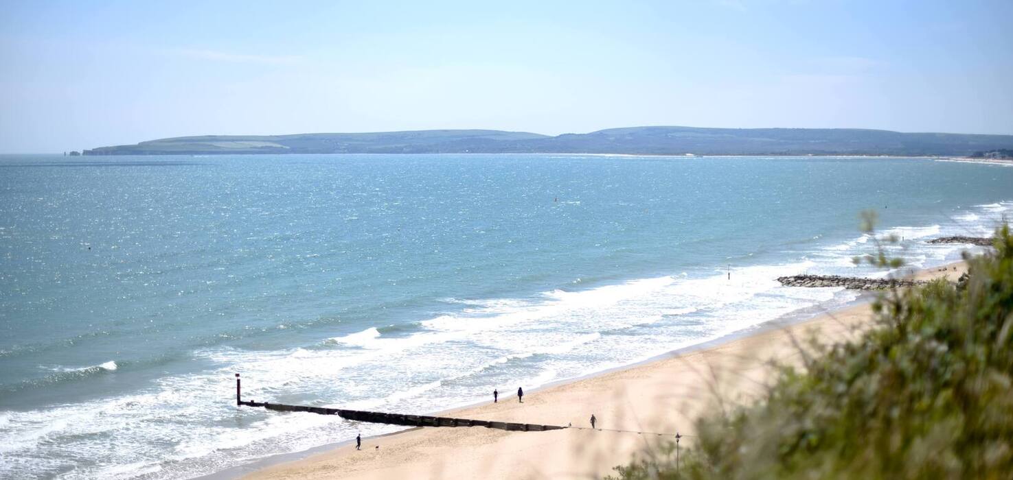 Sandy beachfront with gentle turquoise waters, wooden groynes and distant headland; sunlit shore with people walking.