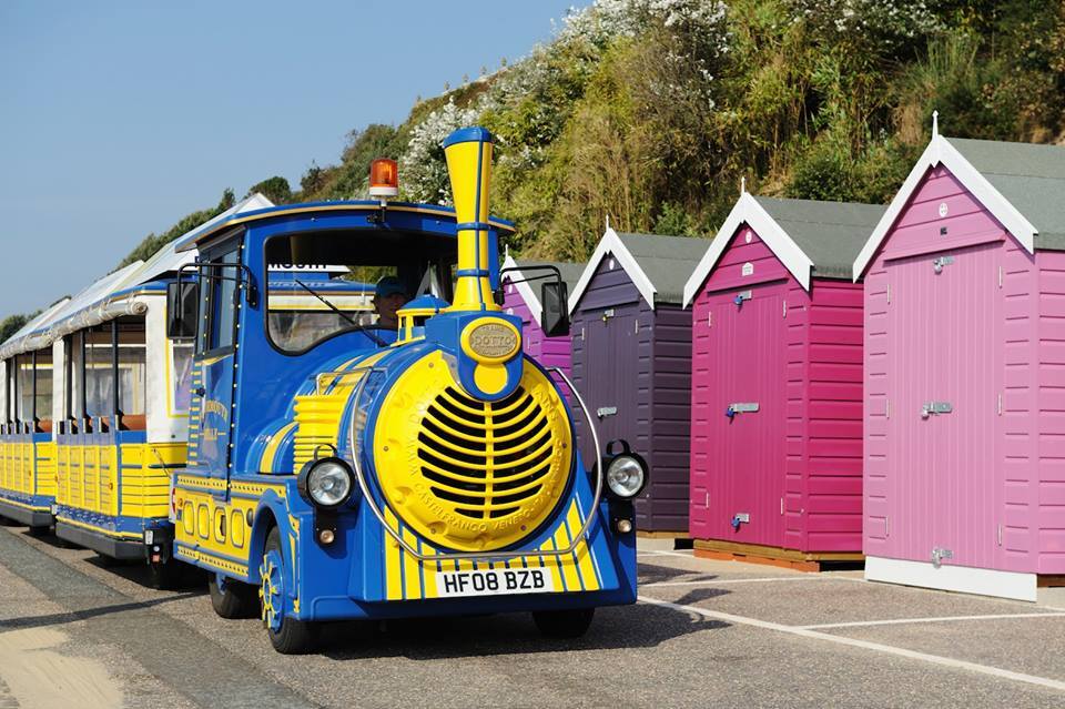 Bright blue-and-yellow tourist train beside pink and purple beach huts on a sunny seaside promenade for family rides