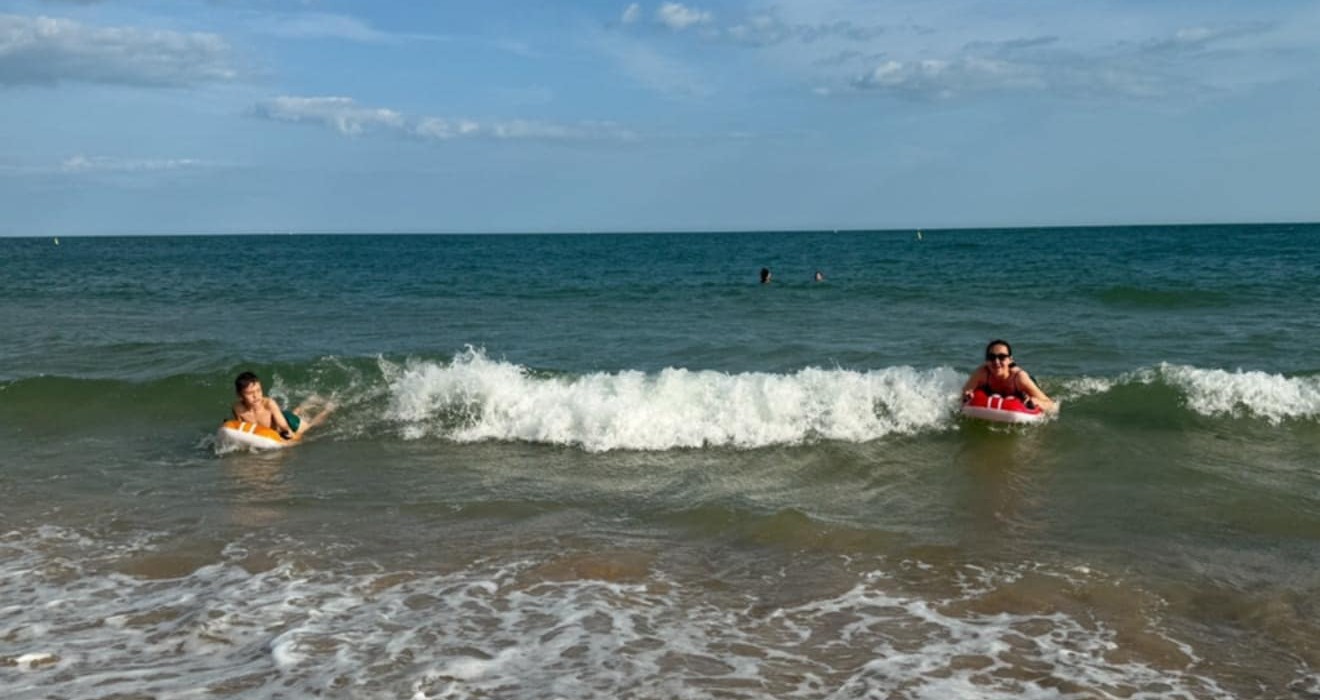 Sunny beachfront with gentle waves and two guests riding inflatable bodyboards near shore