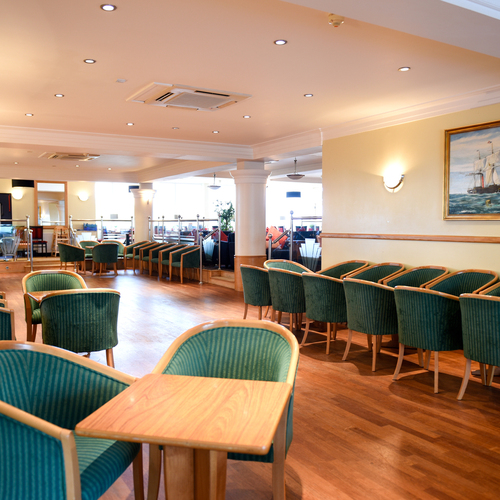 Spacious hotel lounge with rows of green upholstered chairs, wooden tables, warm lighting and a nautical painting.
