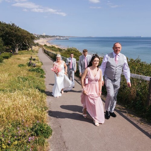 Bridal party in pastel attire walking a cliff-top seaside path with ocean view—romantic beachfront wedding vibe