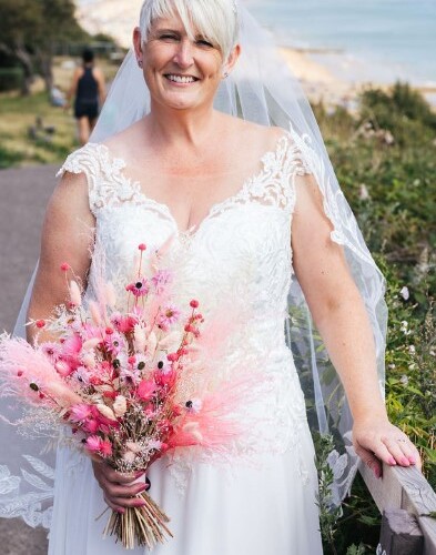 Smiling bride in lace gown and veil holding pink dried-flower bouquet on a seaside promenade with ocean view