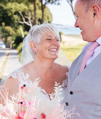 Newlyweds smiling on a tree-lined waterfront path, bride in lace gown with pink bouquet, groom in light-gray suit and pink tie