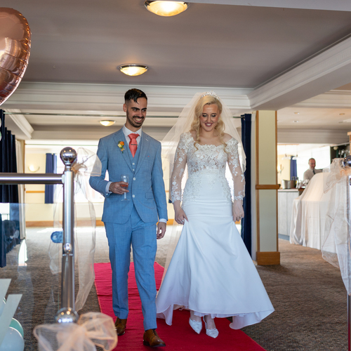 Bride in lace gown and groom in light-blue suit walk a red carpet in a hotel function room; heart balloon with date.