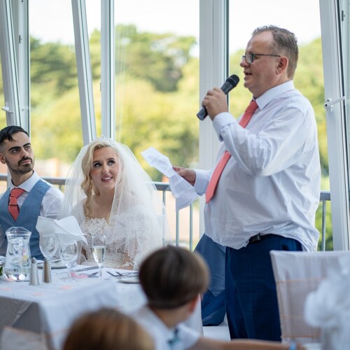 Bride and groom at head table listening to a man's wedding speech, floral centerpiece, glass windows framing green treetop views