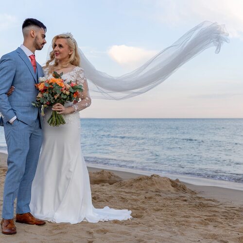 Bride in lace-sleeved gown with flowing veil and orange bouquet beside groom in light-blue suit on beachfront at sunset
