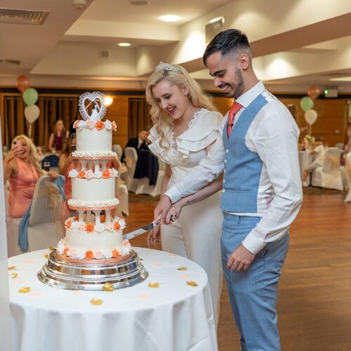 Bride in white gown and groom in blue suit cut a three-tier cake with peach roses in a hotel ballroom