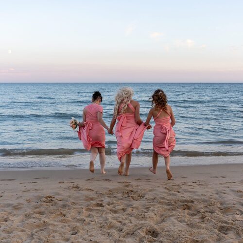 Three bridesmaids in coral-pink dresses holding hands and a bouquet, running barefoot into the calm ocean at sunset.