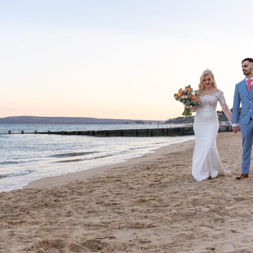 Bride in lace-sleeved gown holding orange bouquet and groom in light-blue suit walk hand-in-hand on sandy beach at sunset.