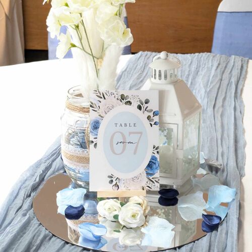Wedding table centerpiece: white lantern, mason jar of cream blooms, mirrored base, scattered blue petals and 'Table 07' card