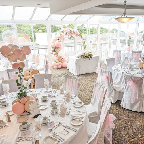 Sunlit glass event room set for a wedding: round tables with white linens, pink balloon centerpieces, arch and green views.