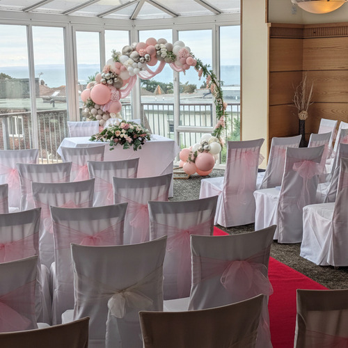 Bright ocean-view ceremony room with floor-to-ceiling windows, white chairs tied with pink bows, floral balloon arch and red aisle.