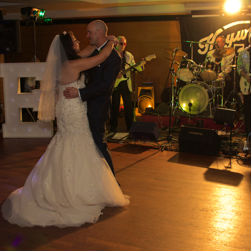 Bride and groom share a first dance on a wooden floor beside illuminated 'LOVE' letters, live band in sparkling jackets.