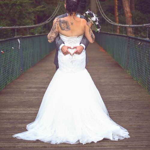 Bride and groom embraced on wooden bridge in forest; bride in strapless gown with tattoos holds bouquet, groom forms heart with hands