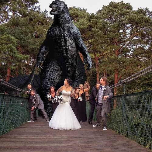 Bridal party on a forest suspension bridge, bride in white gown poses as a giant Godzilla statue looms behind.