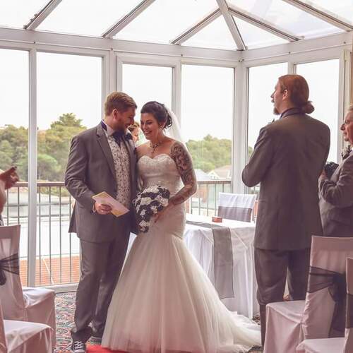 Newlyweds smiling during intimate waterfront ceremony in hotel glass conservatory; bride in gown with bouquet, guests applauding.
