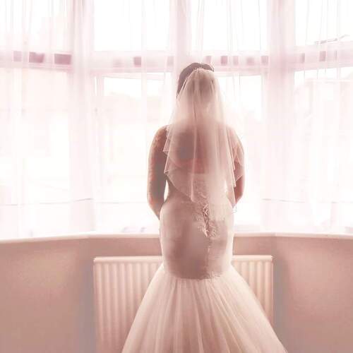 Bride in wedding gown and veil standing at sunlit bay window of a suite, sheer curtains and romantic, airy ambiance.