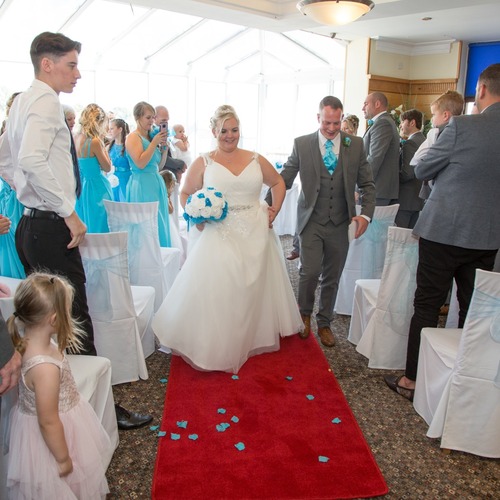 Bride in white gown and groom in gray suit walk down red aisle in a hotel ceremony room, guests and bridesmaids in turquoise dresses