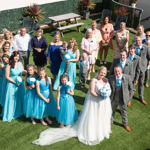 Wedding party on sunny hotel lawn terrace; bride in white gown, groom and groomsmen in grey with turquoise ties, bridesmaids and girls in turquoise.