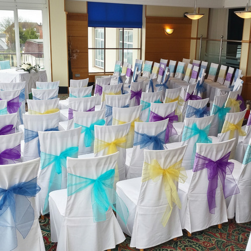 Hotel ceremony room: rows of white-covered chairs tied with teal, purple and yellow bows, front table by glass wall.