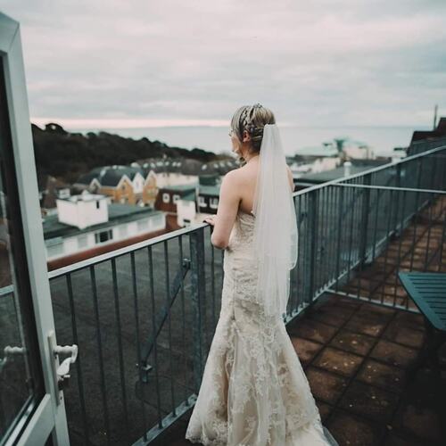 Bride in lace gown and veil on hotel rooftop balcony overlooking coastal town and sea at dusk