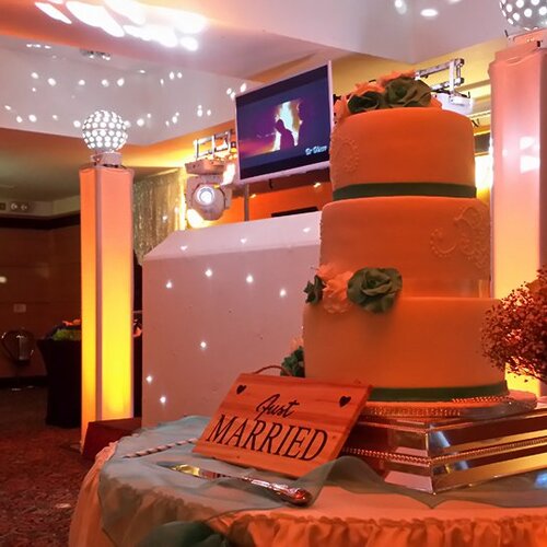 Three-tier wedding cake with green sugar roses and 'Just Married' sign on a lit reception table in warm orange event room.