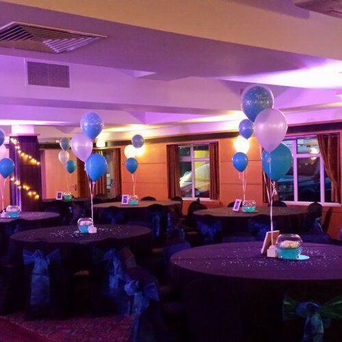 Hotel banquet room with round tables, teal and white balloon centerpieces, chair sashes, purple uplighting and string lights